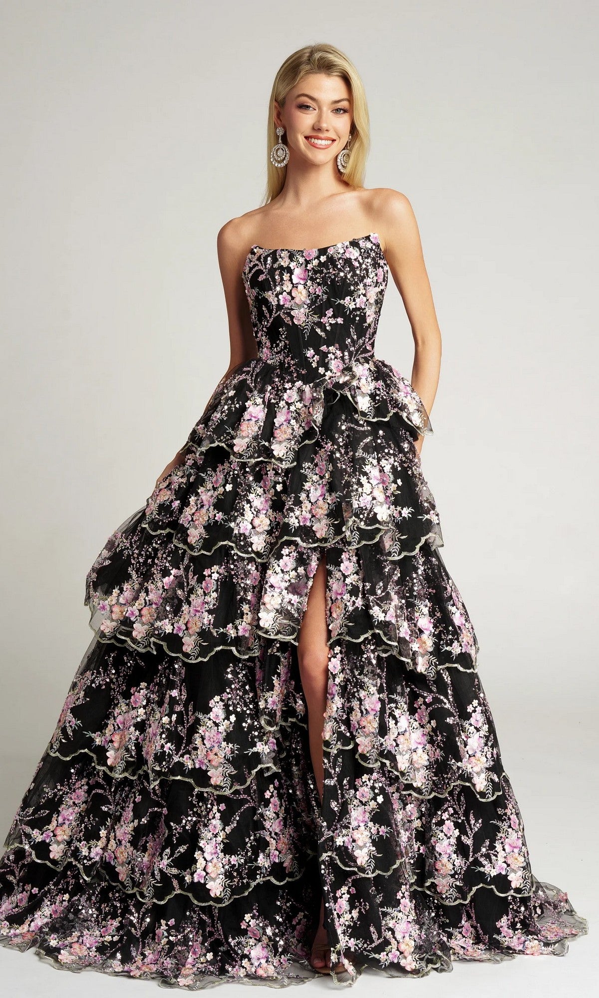 A woman wearing the Portia and Scarlett PS26051 Strapless Floral Ball Gown in black, with embroidered pink and white floral applique, tiered layers, and a front slit, stands and smiles.